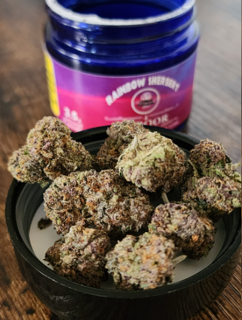 Several cannabis buds rest on a black lid in the foreground, with a purple and pink-labeled container marked "Rainbow Sherbert" open in the background on a wooden surface.