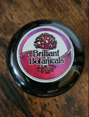 A round container labeled "Brilliant Botanicals" with a stylized tree logo on a pink and white background, placed on a wooden surface.