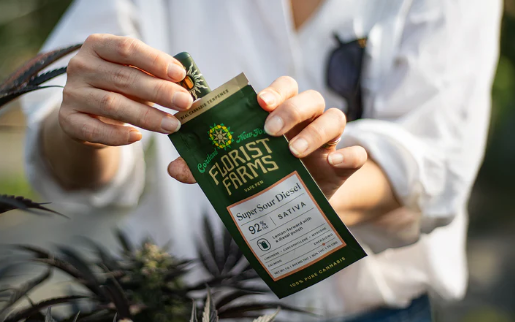 A person in a white shirt holds a green Florist Farms cannabis product package labeled “Super Sour Diesel” near a cannabis plant.
