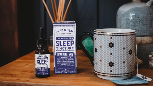 A bottle and box of Head & Heal Sleep Tincture are displayed on a wooden surface next to a patterned mug with tea, suggesting a calming bedtime routine.