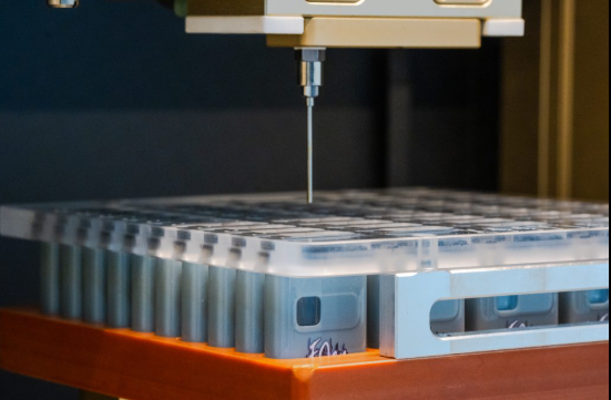 A close-up of a laboratory robotic arm with a thin needle above a tray of sample containers, preparing for automated liquid handling or analysis.