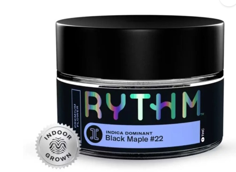 A glass jar labeled "RYTHM" with a black lid contains cannabis flower. The label reads "Indica Dominant Black Maple #22" and "Indoor Grown" with a silver indoor grown seal visible.