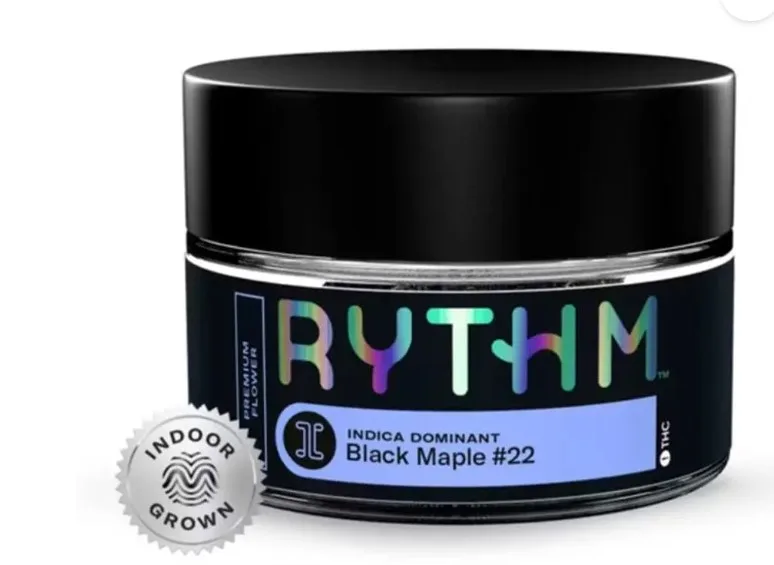 A glass jar labeled "RYTHM" with a black lid contains cannabis flower. The label reads "Indica Dominant Black Maple #22" and "Indoor Grown" with a silver indoor grown seal visible.