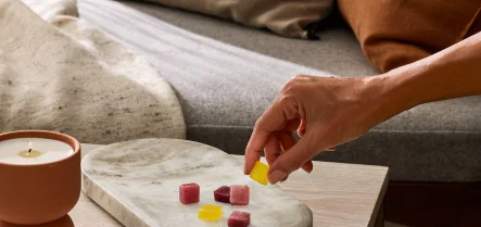 A hand picks up a yellow gummy from a marble tray with assorted gummies and a lit candle on the table.