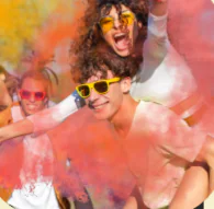 People wearing sunglasses enjoy a color festival, smiling as bright powder fills the vibrant air around them.
