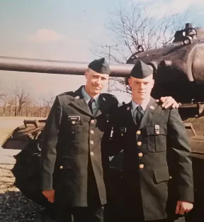 Two men in military uniforms smile in front of a tank, with one man’s arm around the other.