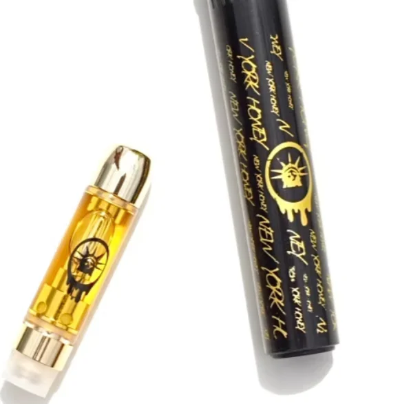A vape cartridge with golden liquid sits beside a case labeled “NEW YORK” featuring the Statue of Liberty.
